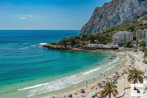 A coastal beach with clear turquoise waters, a rocky outcrop, and a row of buildings with balconies overlooking the sea.