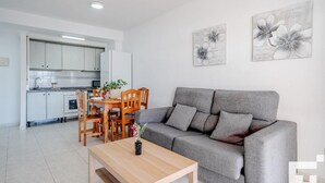 A modern living room with a grey sofa, wooden dining table, and chairs. There is a kitchen in the background with white cabinets and a built-in oven. Two floral wall art pieces are displayed above the sofa. - Zafiro 35C - Grupo Turis - Calpe, Costa Blanca (Calpe)