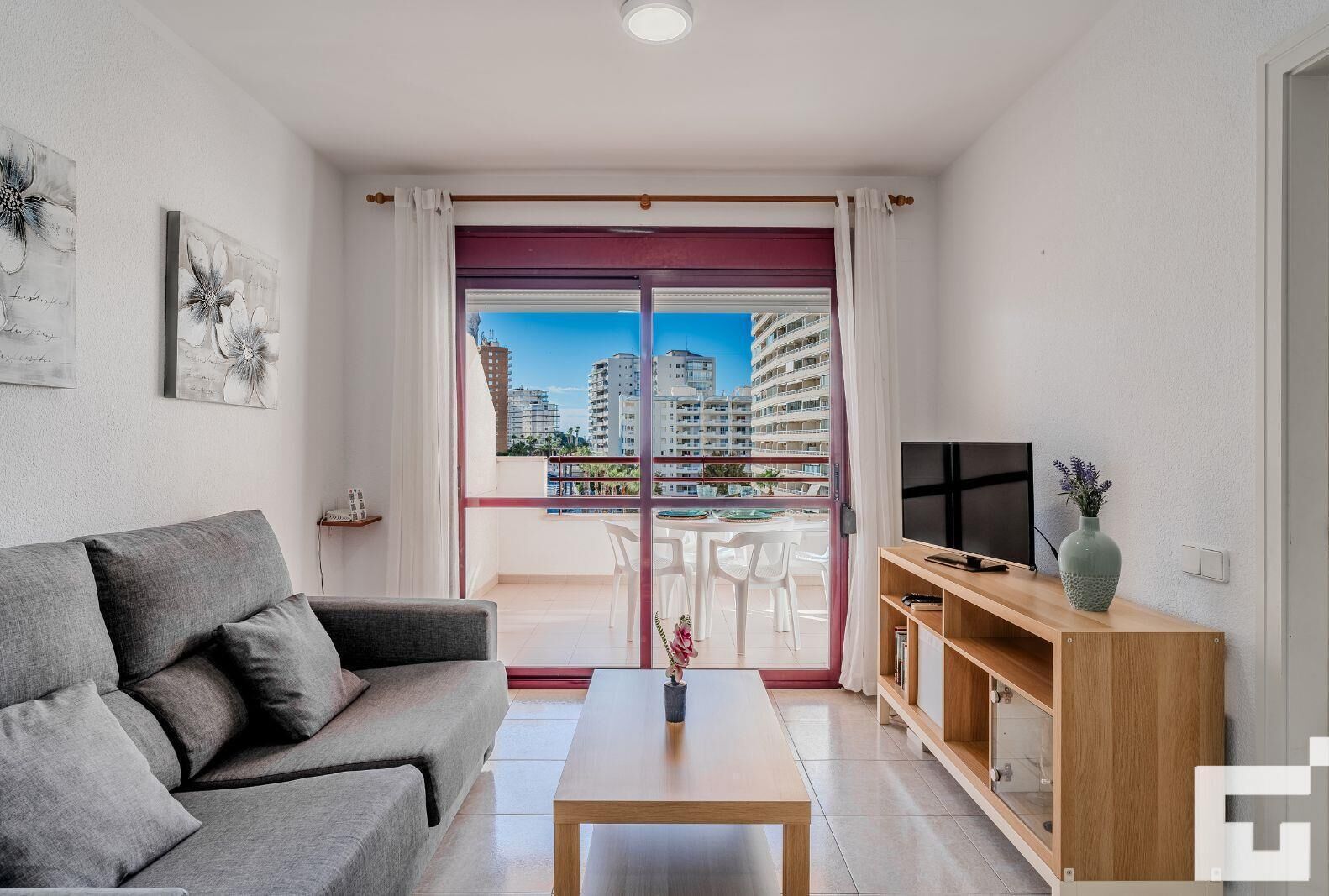 A modern living room with a grey sofa, a wooden coffee table, a TV on a wooden stand, and a balcony with a city view.