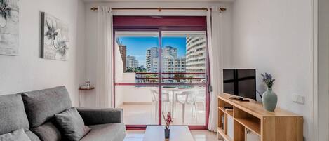 A modern living room with a grey sofa, a wooden coffee table, a TV on a wooden stand, and a balcony with a city view.