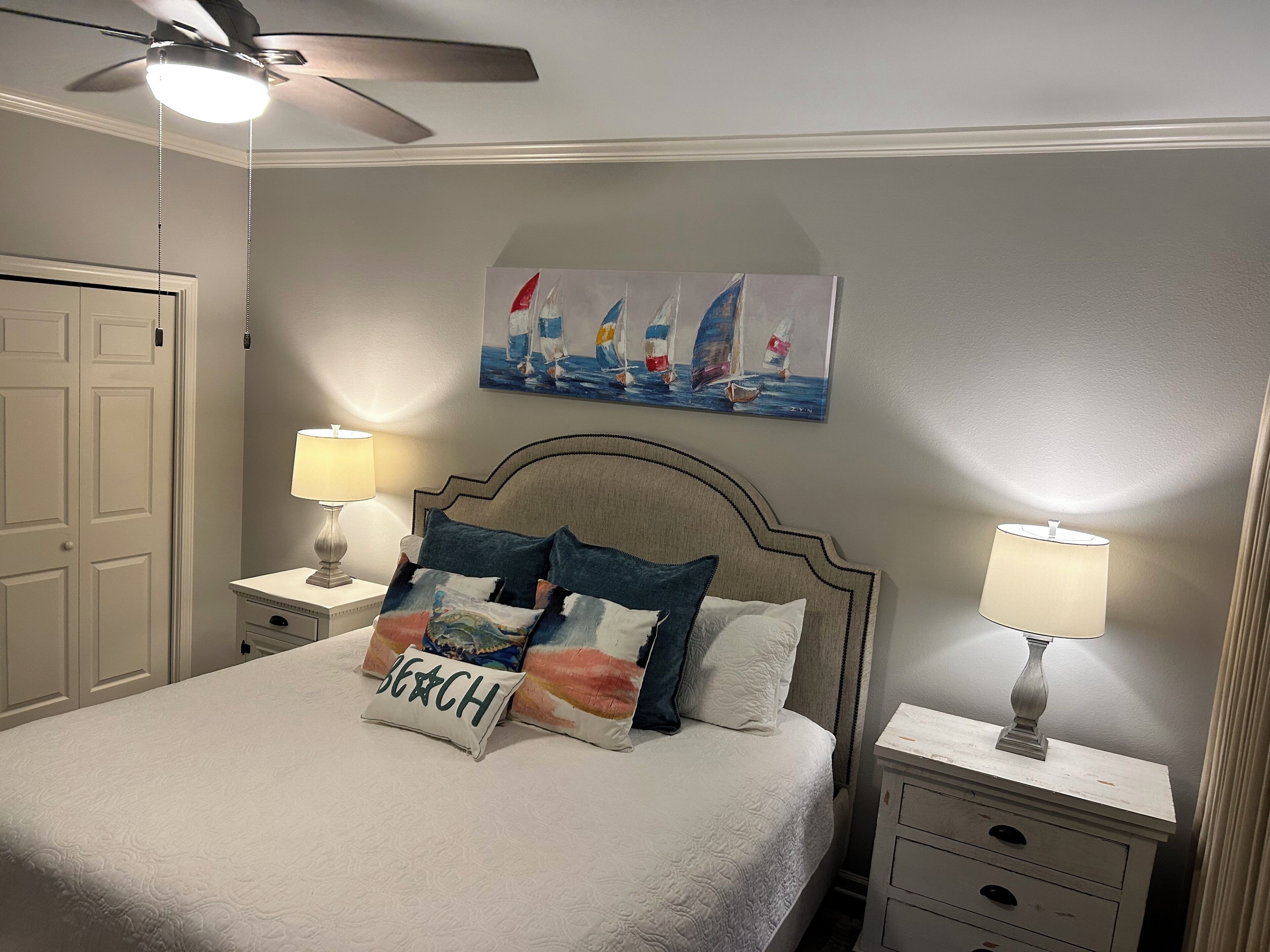 A neatly arranged bedroom with a bed, two bedside tables with lamps, a ceiling fan, a painting of sailboats, and a door.