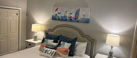 A neatly arranged bedroom with a bed, two bedside tables with lamps, a ceiling fan, a painting of sailboats, and a door.