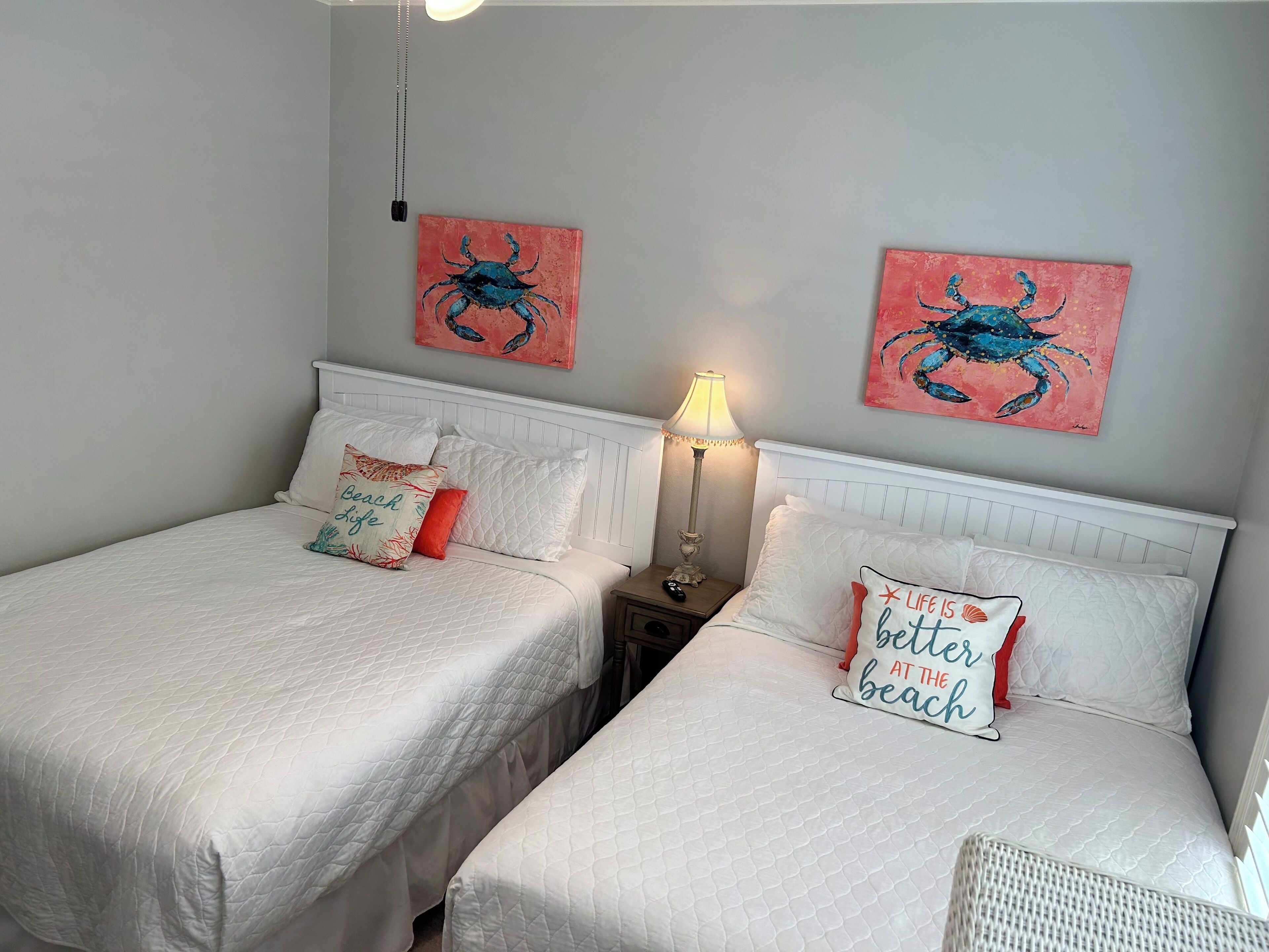 Een slaapkamer met twee bedden, elk voorzien van witte lakens en decoratieve kussens, een nachtkastje met een lamp, en twee aan de muur bevestigde schilderijen van krabben.
