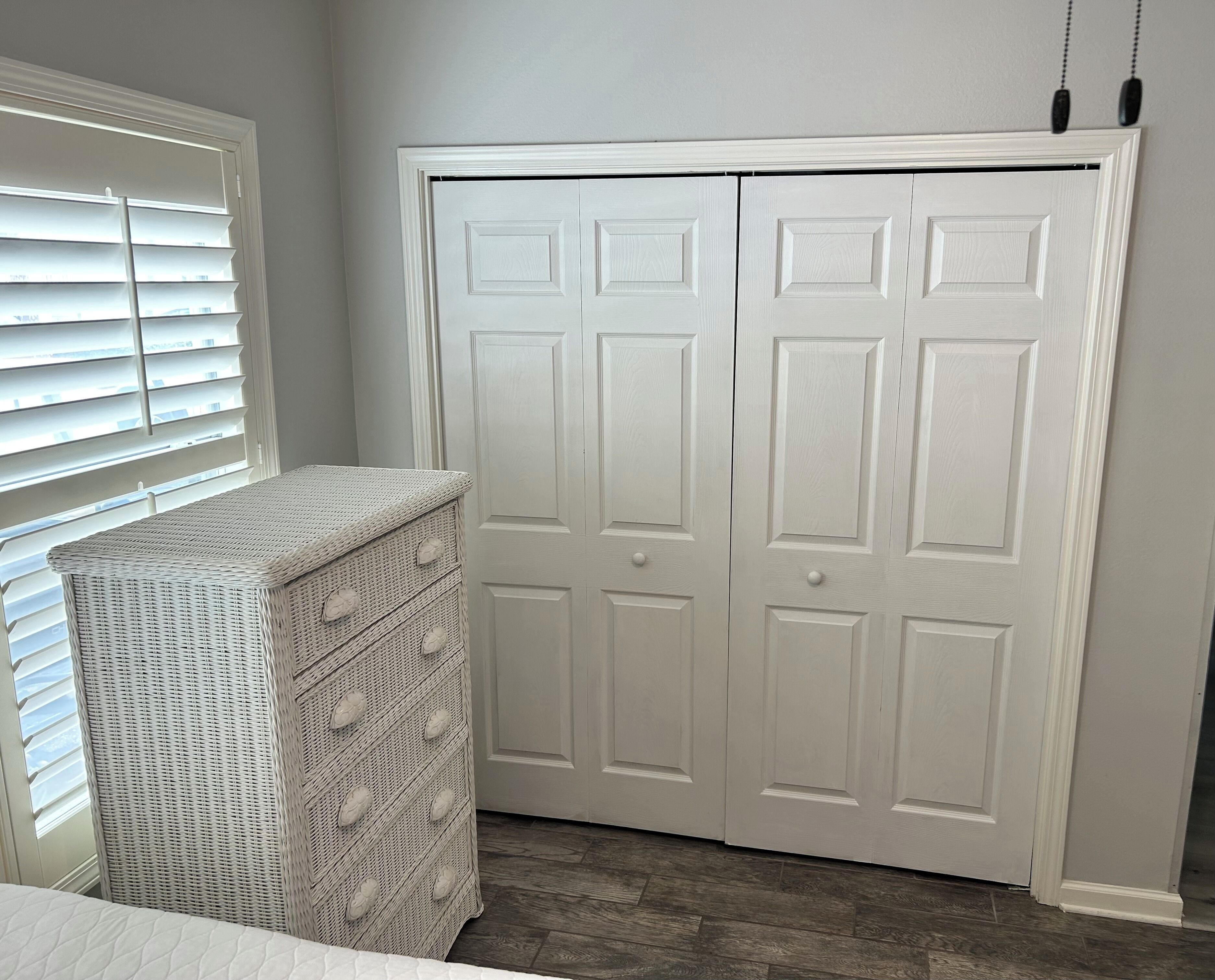 A room with a white wardrobe, a wicker chest of drawers, and a window with blinds.