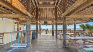 Une terrasse extérieure en bois, dotée de tables de pique-nique et de poubelles, avec vue sur une plage.