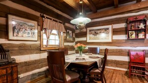 Un comedor rústico con paredes de madera, una mesa de madera y sillas. Hay cuadros en las paredes, una lámpara colgante y un jarrón con flores sobre la mesa.