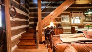 Interior de una cabaña de troncos con una escalera de madera, un sofá de cuero y un comedor con mesa y sillas.
