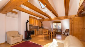Una sala de estar acogedora con un sofá, una alfombra roja, vigas de madera, una cocina con armarios de madera y una zona de comedor con mesa y sillas.