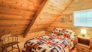 A cozy attic bedroom with a wooden sloped ceiling, a bed with a patterned quilt, a chair, a nightstand, and a window with blinds. - Large Mountain Creek! Cabin in Tellico Plains (Tellico Plains)
