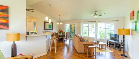 A spacious living area with hardwood flooring, a sofa, a dining table, and a TV. There are ceiling fans, a kitchen area, and large windows providing natural light.