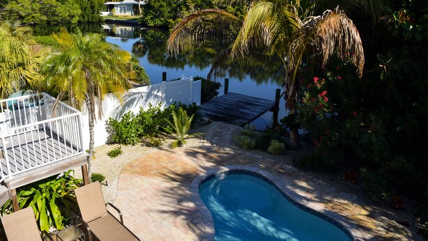Una piscina rodeada de abundante vegetación, una terraza de madera y una valla blanca.