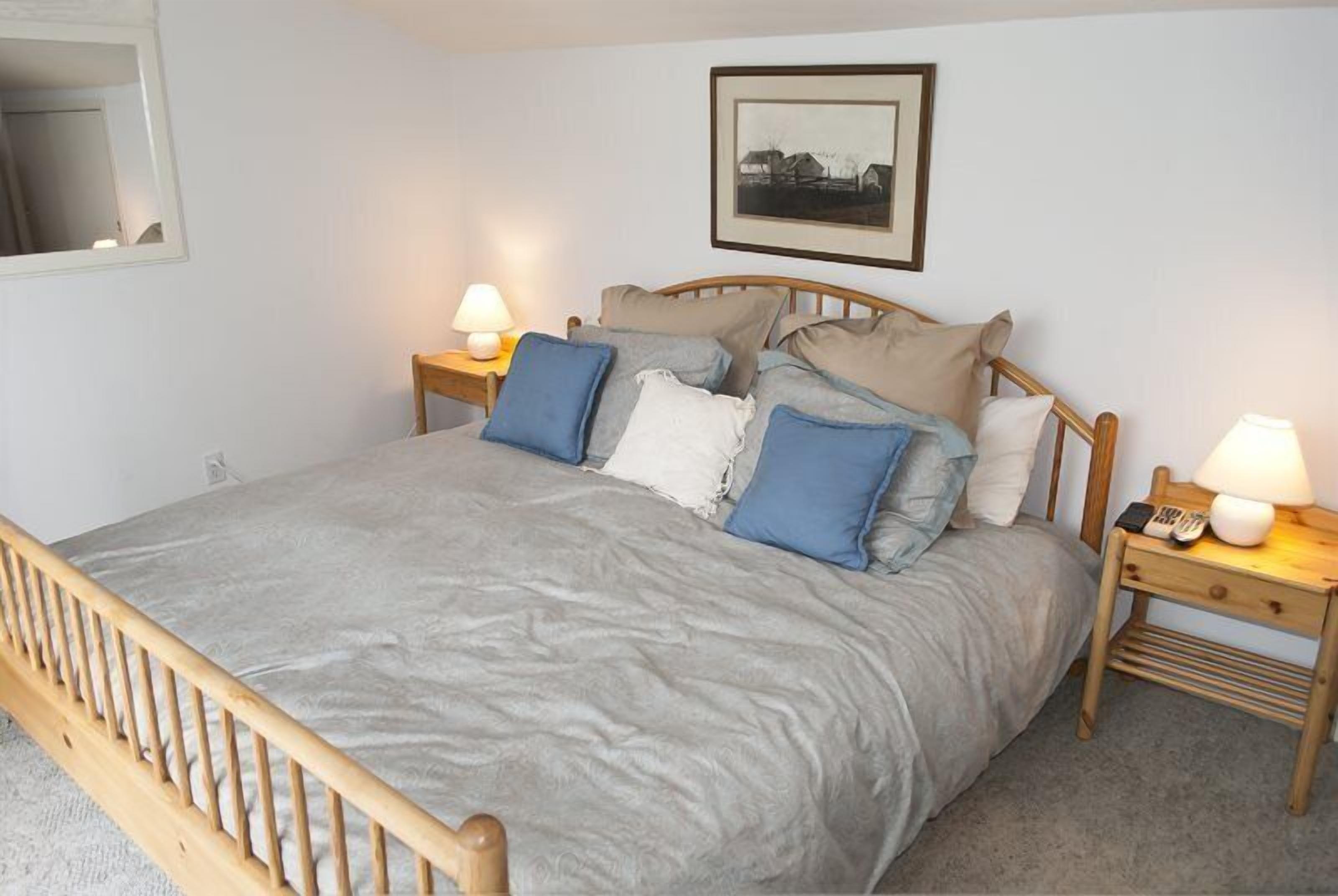 A neatly made bed with a grey bedspread and multiple pillows, two bedside tables with lamps, and a framed picture on the wall.