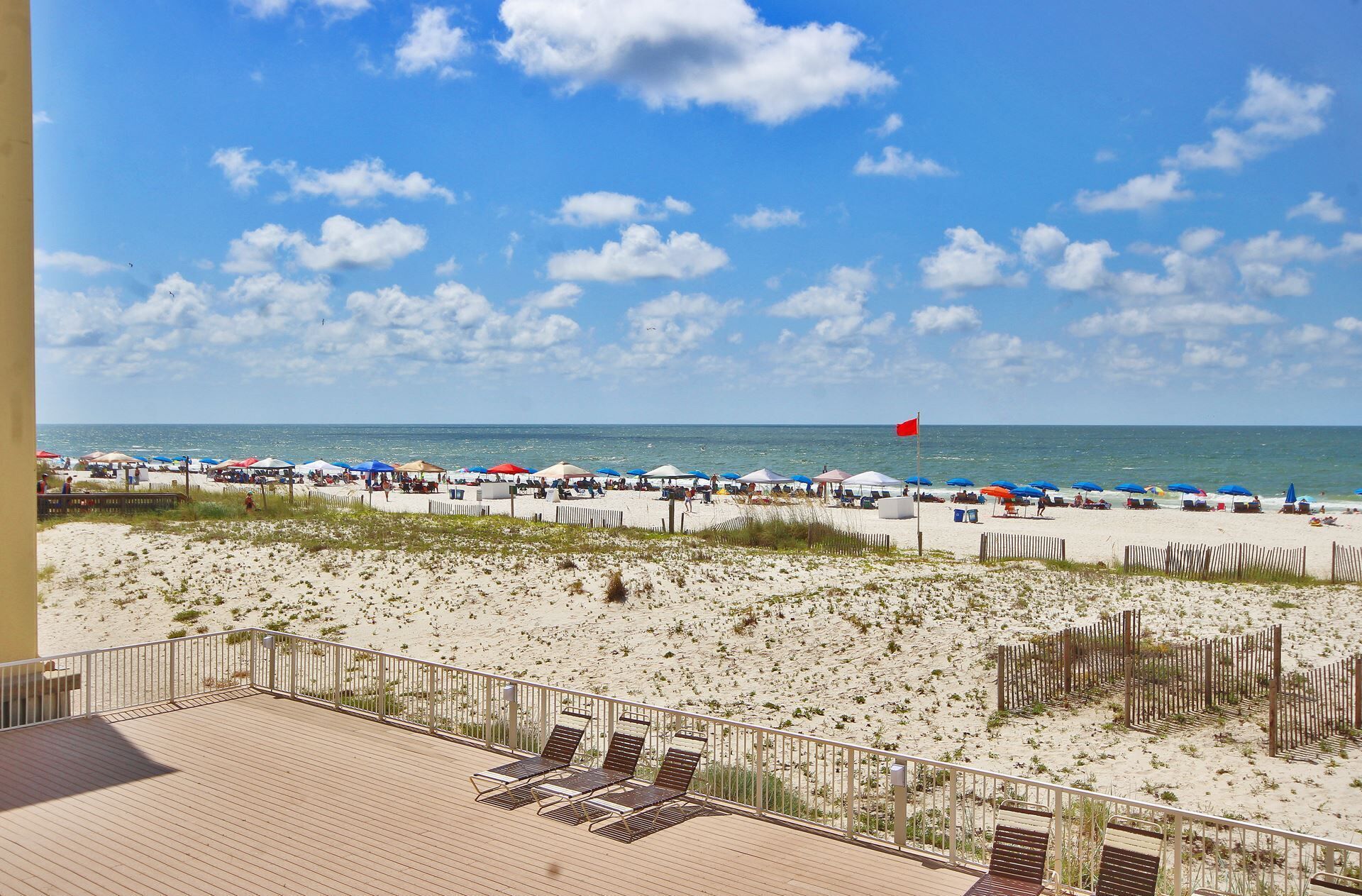 A beach view with a wooden deck and lounge chairs, a sandy beach with umbrellas, and a flag in the distance.