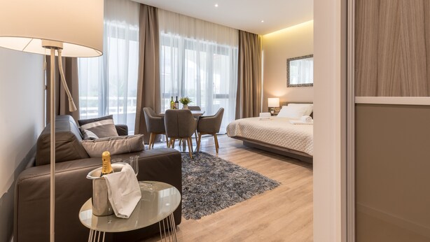 Et moderne hotelværelse med sofa, et rundt bord med champagne, seng, skrivebord og et stort vindue med gardiner.
