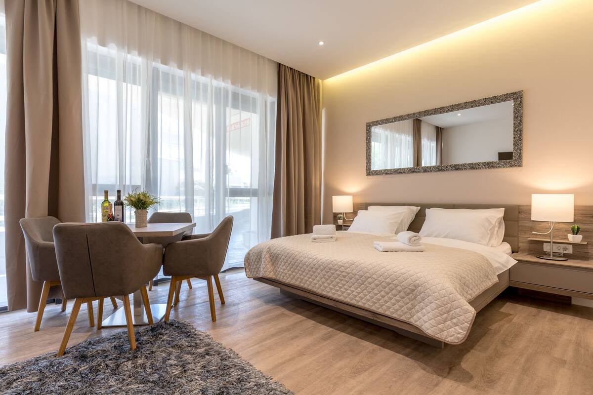 A hotel room with a bed, bedside tables, a desk with chairs, and a large window with curtains.