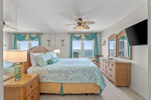 Quarto com cama, penteadeira, mesa de cabeceira, ventilador de teto e vista para o mar.