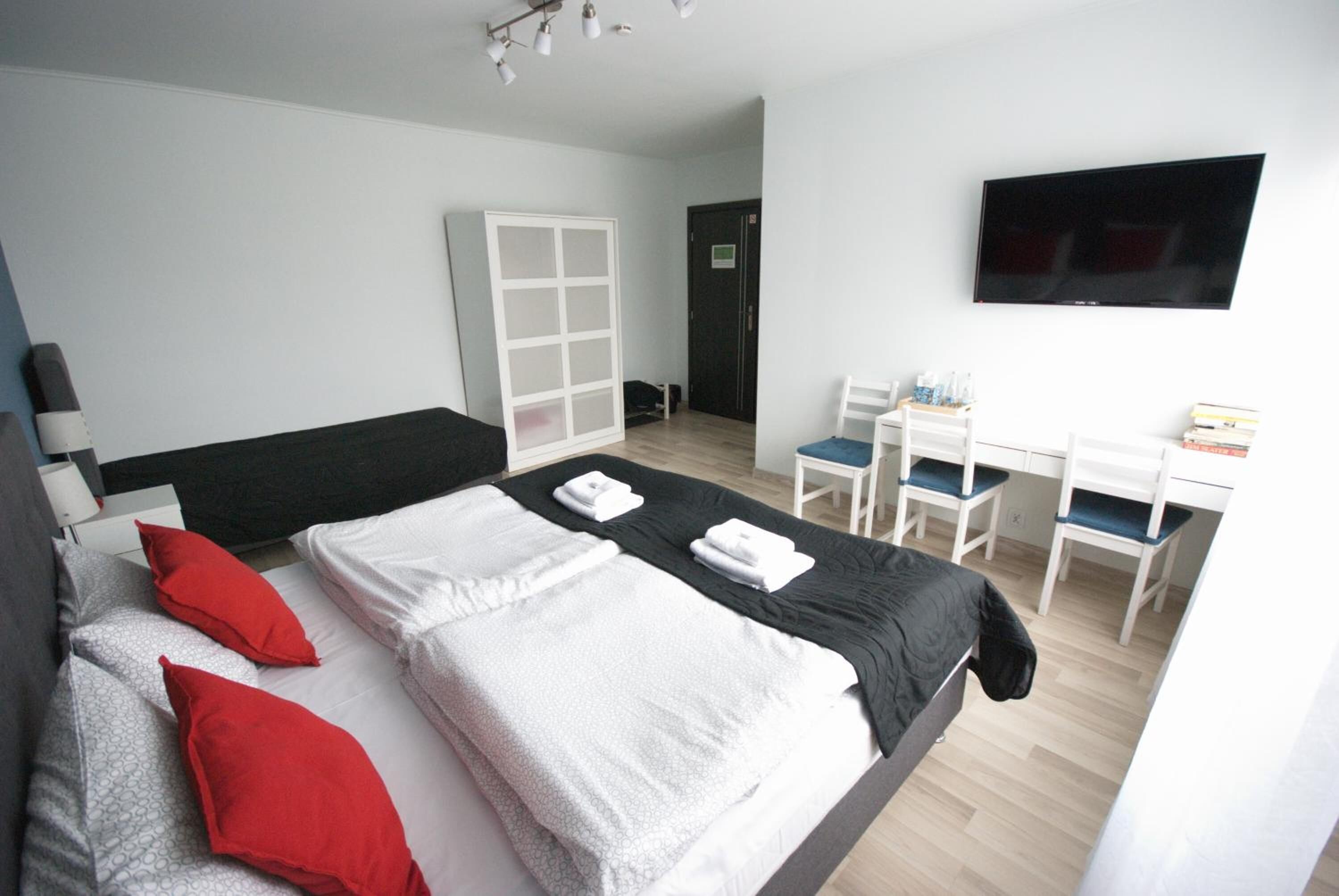 Un dormitorio con cama, dos almohadas rojas, colcha blanco y negro, escritorio con sillas, televisor y armario.