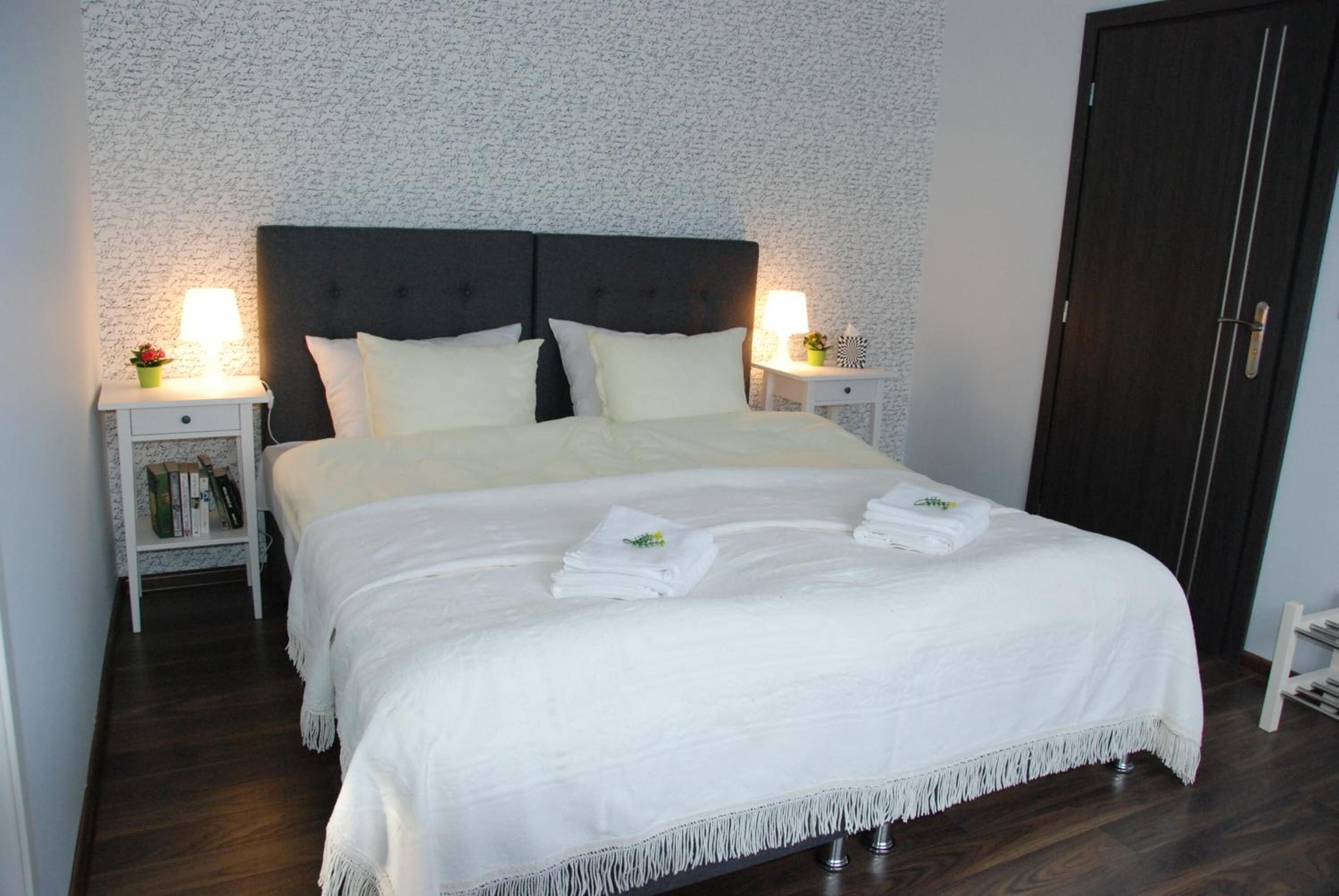 A neatly made bed with white linens and a dark headboard, flanked by bedside tables with lamps and small decorative items.