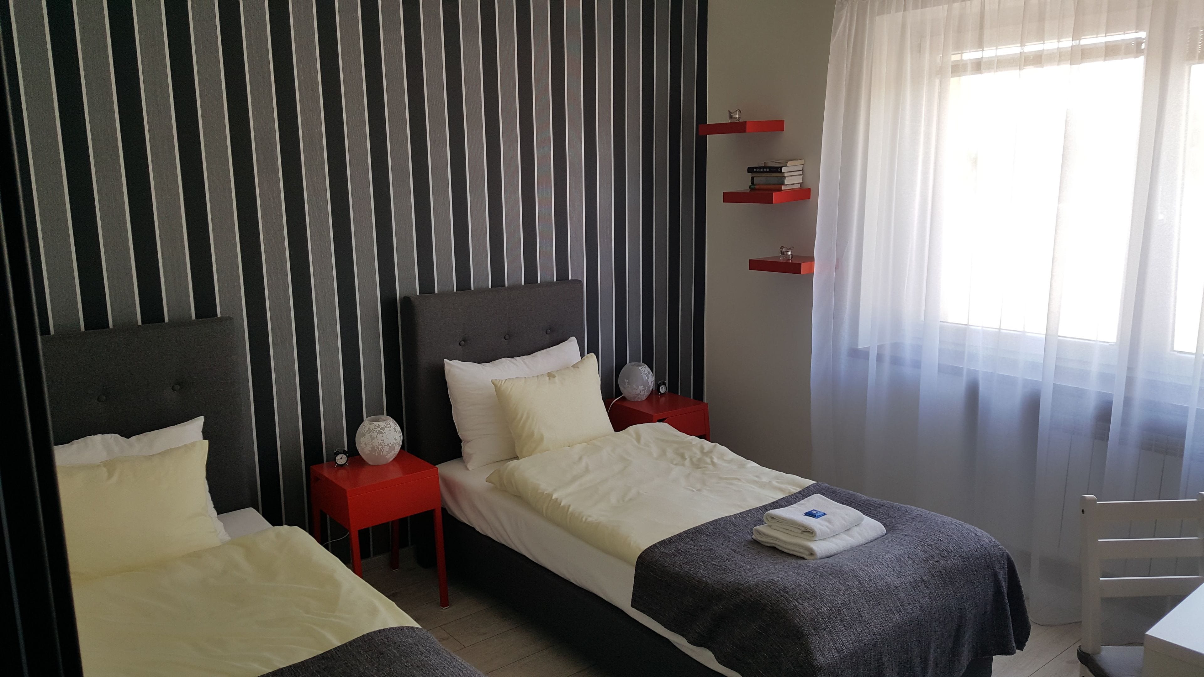 Une chambre d’hôtel avec deux lits, un papier peint à rayures, une table de chevet rouge et une fenêtre avec des rideaux légers.