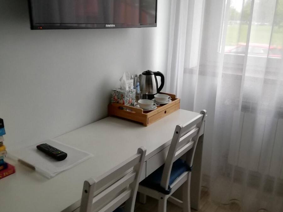 Une petite pièce simple, avec un bureau blanc, deux chaises, un téléviseur fixé au mur et une fenêtre avec des rideaux.