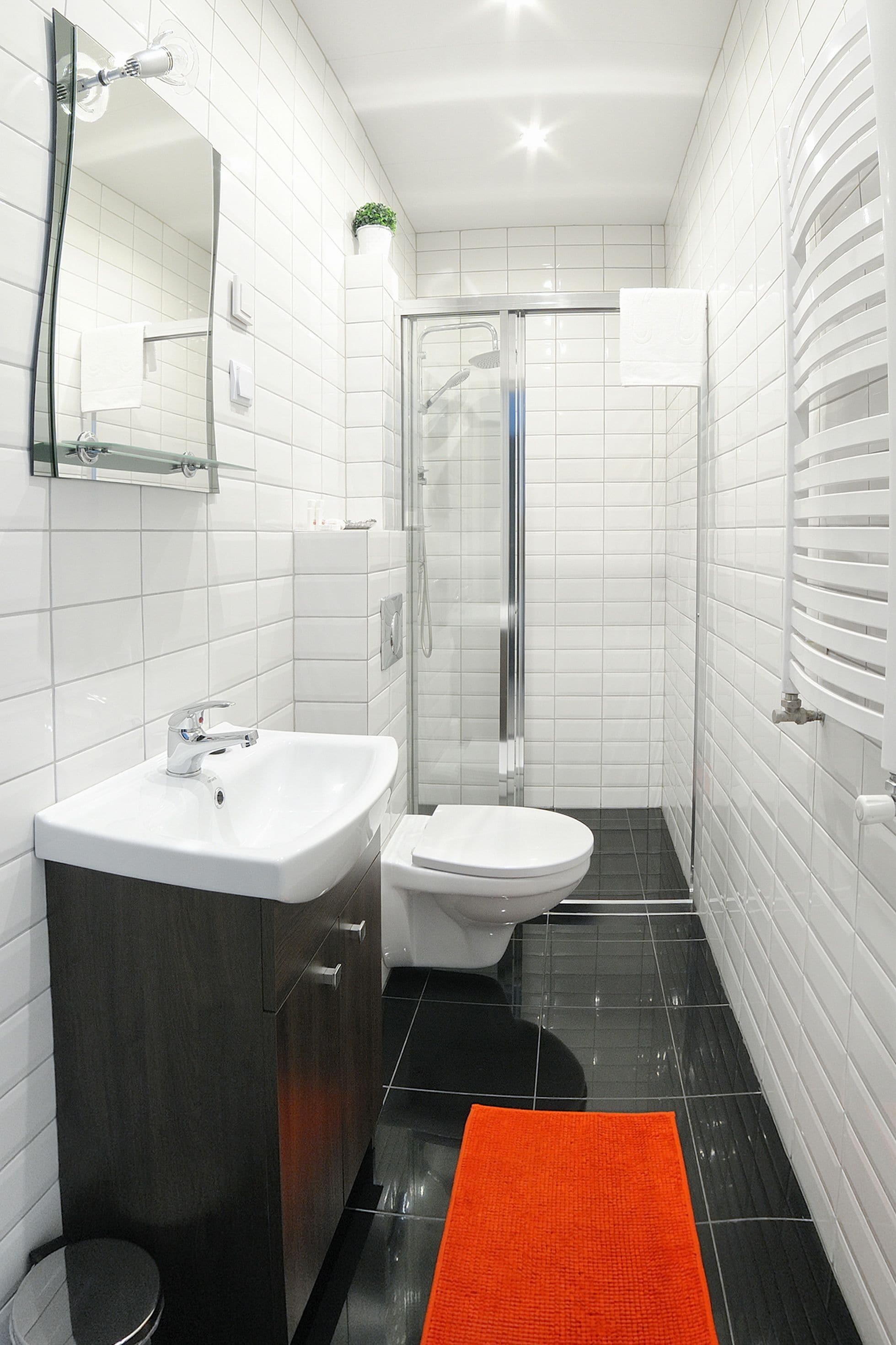 A modern bathroom with white tiles, a black floor, a white sink, a toilet, a glass shower enclosure, and a small potted plant.