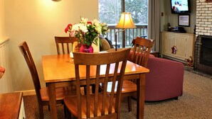 A dining area with a wooden table, chairs, a vase of flowers, a lamp, and a television in the background.