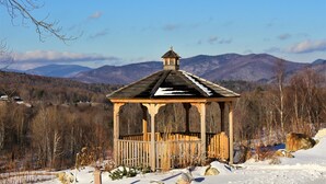 A wooden gazebo with a peaked roof, surrounded by snow-covered ground and mountains in the background.