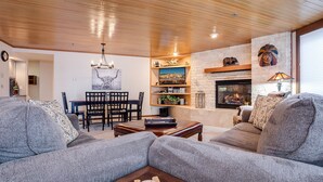 A cozy living room with a fireplace, a dining area with a chandelier, and a large window with blinds.