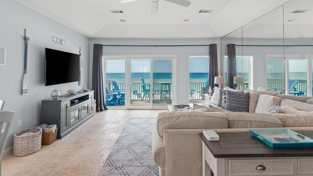 A spacious living room with a large flat-screen TV, a ceiling fan, and a view of the ocean through large sliding doors.
