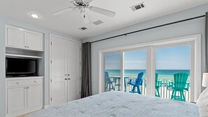 A bedroom with a bed, a ceiling fan, a TV, and a view of the beach through large glass doors.