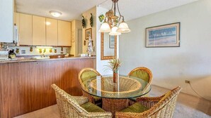 A dining area with a round glass table surrounded by wicker chairs, a kitchen in the background, and a ceiling light fixture. - Fun island vibe! Private lanai, Full Kitchen, Flat Screen TV, Air Conditioning, WiFi- Kamaole Sands (KIHEI)