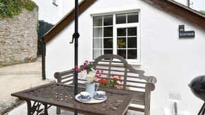 Uma casa de campo branca com uma placa 'Cottage', um banco de madeira, uma mesa de metal com um vaso de flores e uma janela com vidros brancos.