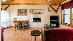 A cozy living room with a stone fireplace, wooden ceiling, a dining table with chairs, a television, and a red curtain. - Alamo Springs- The Barn | Relaxing Retreat (Fredricksburg)