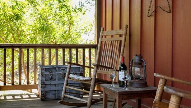 A wooden rocking chair and table on a wooden deck with a rustic appearance, a vintage lantern, and a bottle of wine. - Alamo Springs- The Barn | Relaxing Retreat (Fredricksburg)