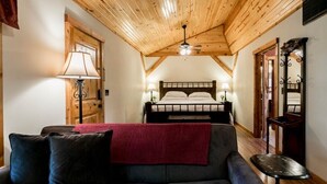 A cozy room with a wooden ceiling, a bed with white linens, a dark sofa with a red throw, and a small table. - Alamo Springs- The Barn | Relaxing Retreat (Fredricksburg)