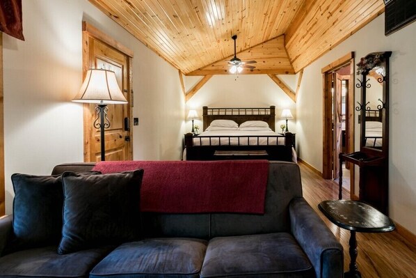 Une pièce chaleureuse avec un plafond en bois, un lit recouvert de draps blancs, un canapé sombre agrémenté d’un plaid rouge et une petite table.