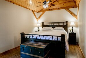 Une chambre avec un plafond en bois, un lit recouvert de linge de lit blanc, deux lampes de chevet et une valise ancienne posée sur le sol.