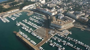 An aerial view of a marina with numerous boats docked, surrounded by residential buildings and a hilly landscape in the background. - HOUSE LA FLOR (L'Escala)