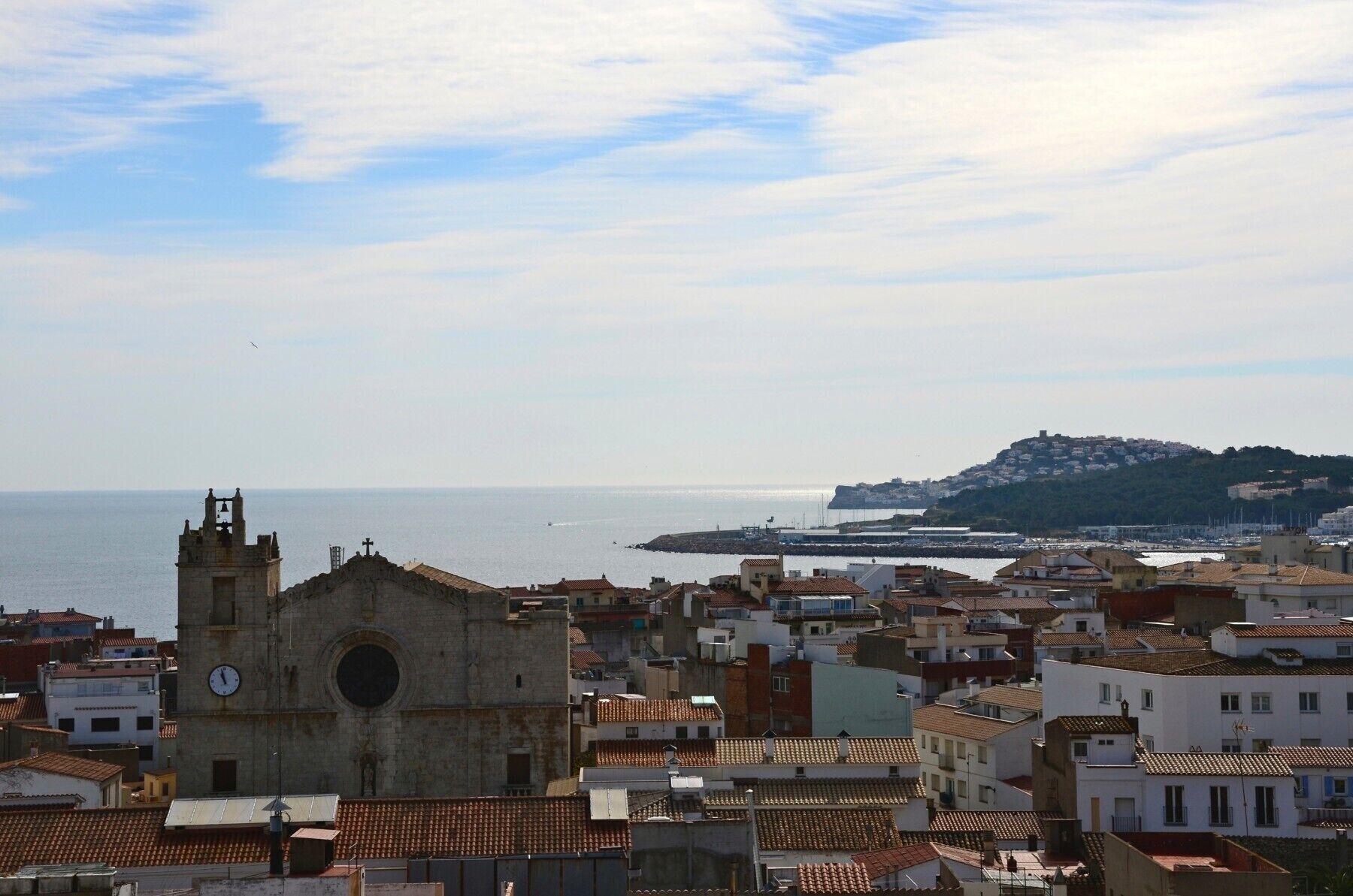 Un paisaje urbano costero con una imponente torre de iglesia, una colina con edificios y un puerto deportivo.