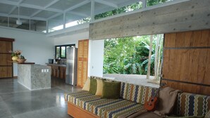 Una sala de estar moderna con un sofá, una ventana con vistas a la vegetación y una cocina al fondo.