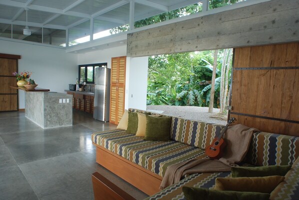 A modern living room with a sofa, a window overlooking greenery, and a kitchen area in the background. - Amazing Luxurious Pool House With Big Views and Privacy Best Beach Playa Hermosa (Hermosa de Cobano)