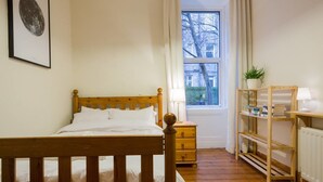 A bedroom with a wooden bed, a nightstand, a window with a view of buildings, a shelf with books, and a framed picture on the wall.