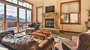 A spacious living room with a stone fireplace, leather sofas, and a large window offering a view of the outdoors. - Crested Butte Townhome w/ Views - Steps to Lifts! (Mt. Crested Butte)