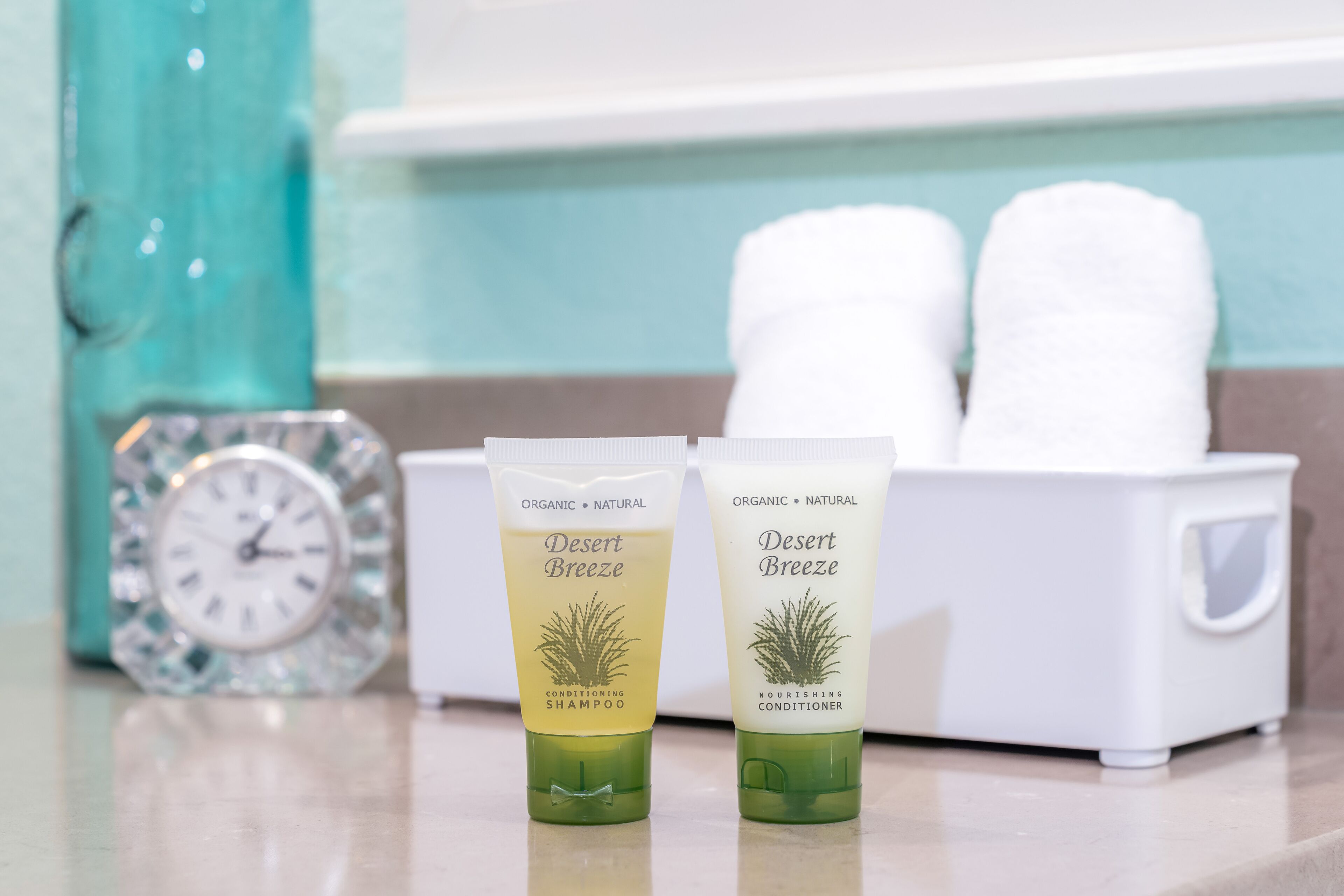 Two tubes of Desert Breeze organic shampoo and conditioner on a bathroom counter with a clock and towels in the background.