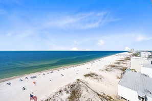 A beach with white sand, clear blue water, and a few buildings in the background.