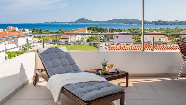 A balcony with a sun lounger, a small table with fruit and a drink, and a view of the sea and buildings. - Apartments Arausa with Pool (Vodice)