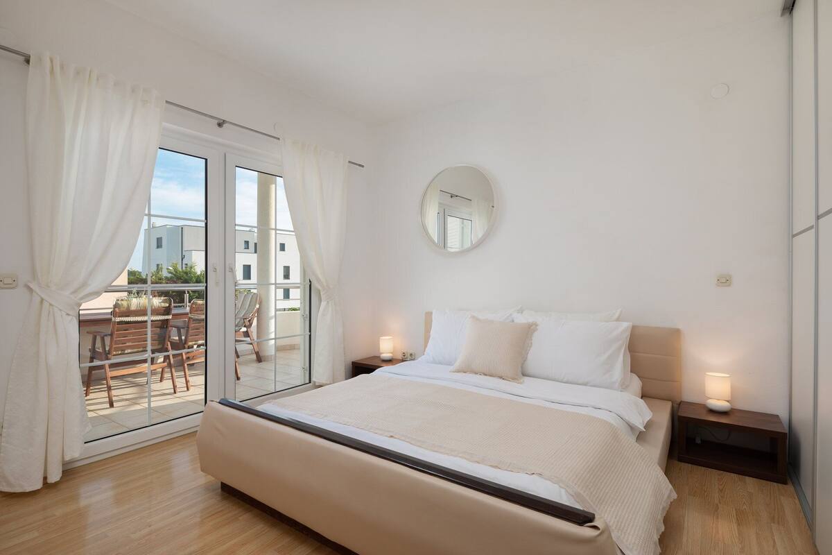 A bedroom with a large bed, a round mirror, and a balcony with chairs.