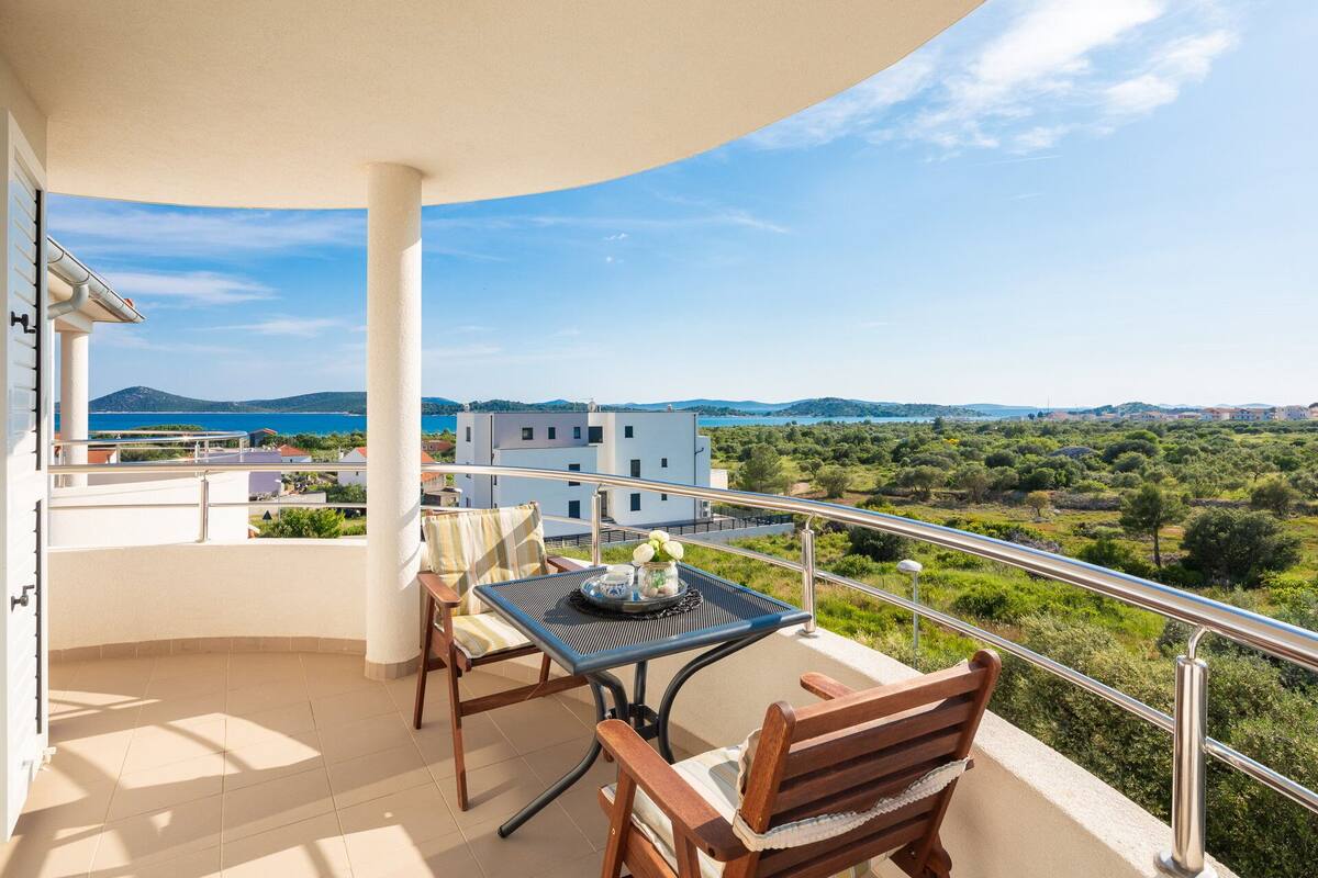 A balcony with a table and chairs, overlooking a scenic landscape with buildings and greenery.