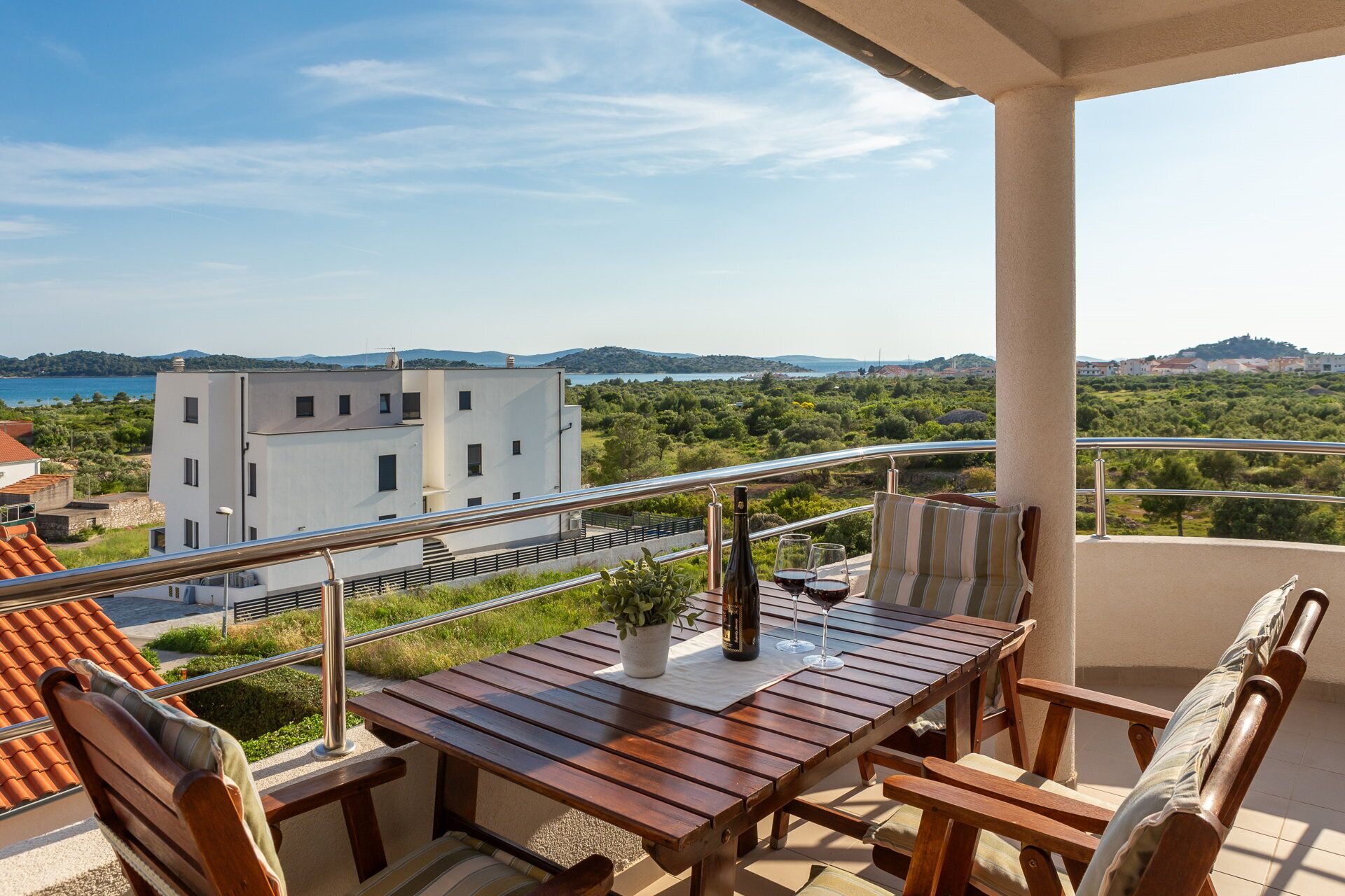 A balcony with a wooden table and chairs, a bottle of wine, and glasses. The view includes a body of water and buildings in the distance.