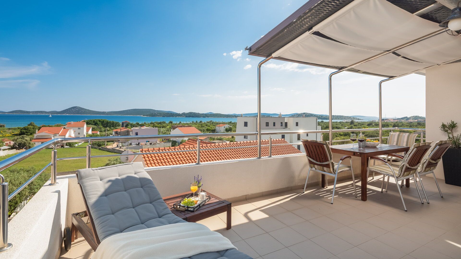 A balcony with a sofa, dining table, and chairs, overlooking a coastal view with houses and a clear sky.